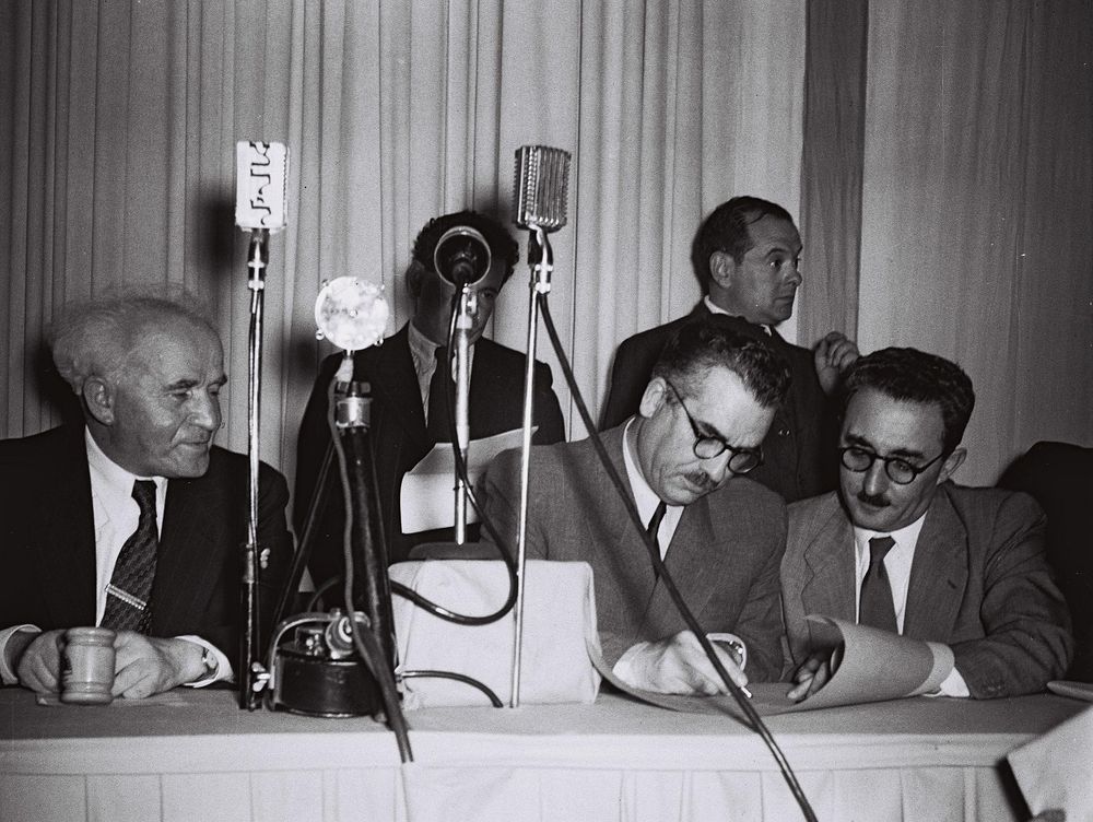DAVID BEN GURION WATCHING MOSHE SHAPIRA SIGNING THE SCROLL OF THE DECLARATION OF INDEPENDENCE HELD BY MOSHE SHARRET DURING THE CEREMONY IN THE TEL AVIV MUSEUM.