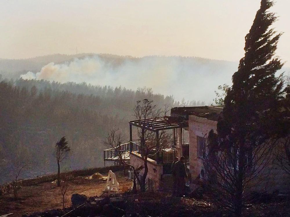 Personnel survey the remains of a house near Nataf as a forest fire burns in the background