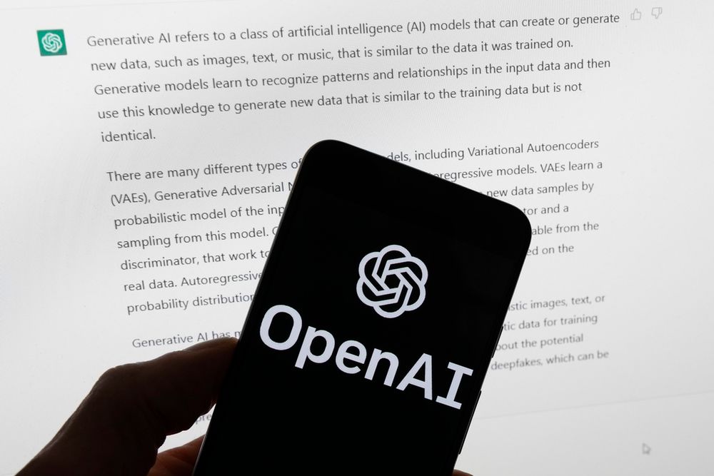 The OpenAI logo is seen on a mobile phone in front of a computer screen which displays output from ChatGPT.