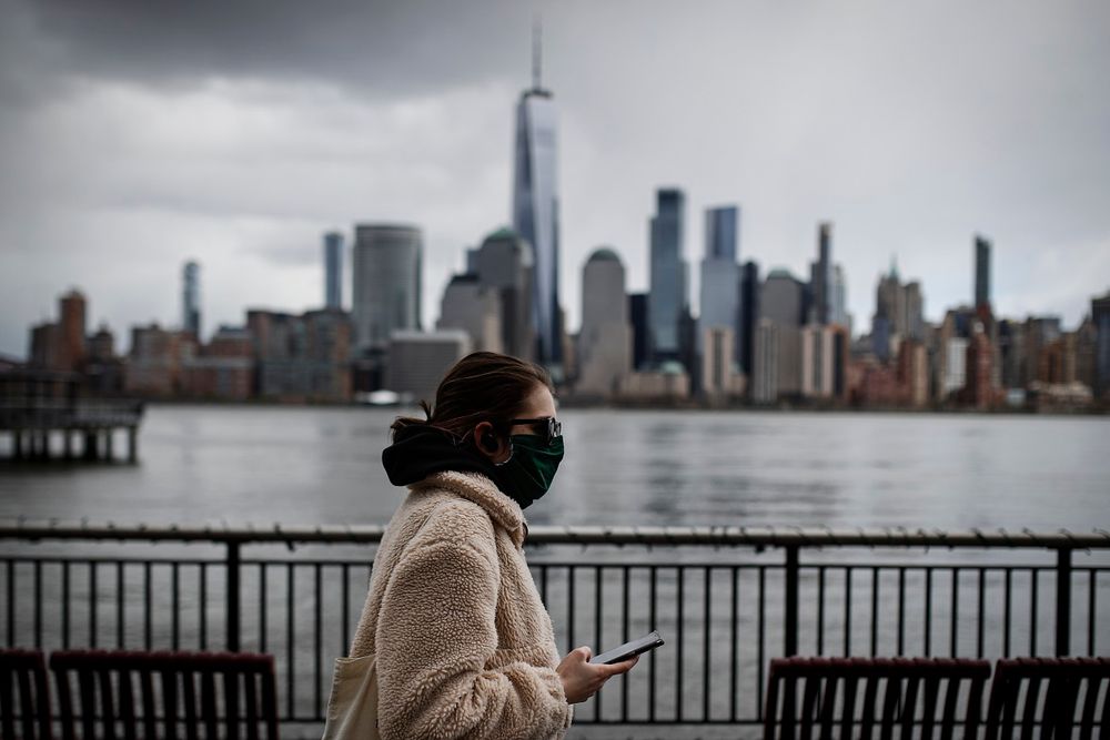 The downtown New York City skyline looms over a pedestrian wearing a mask due to COVID-19 concerns, in Jersey City, N.J on  April 10, 2020.