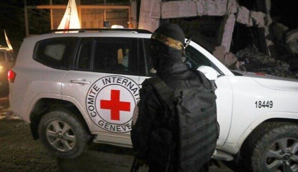 An armed man wearing the uniform of the al-Qassam Brigades, the military wing of Hamas, stands guard as Red Cross vehicles carrying coffins containing the bodies of four deceased hostages leave a warehouse for Israel in Gaza City on Tuesday, October 14, 2025.