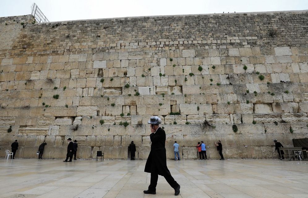 An ultra-Orthodox Jewish man wearing a plastic-covered hat and speaking on a phone walks past people praying at the nearly deserted Western Wall in Jerusalem on March 12, 2020.