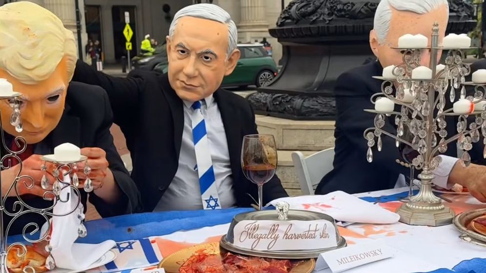 Performers donning the masks of Trump and Netanyahu drink mock blood from wine glasses at a table covered with a blood-spattered Israeli flag.