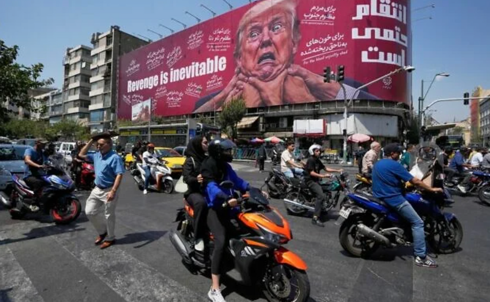 AP Photo/Vahid Salemi Des motocyclistes passent devant une affiche représentant le président américain Donald Trump, les mains autour du cou, sous l’inscription « La vengeance est inévitable », à Téhéran, en Iran, le 24 août 2026.