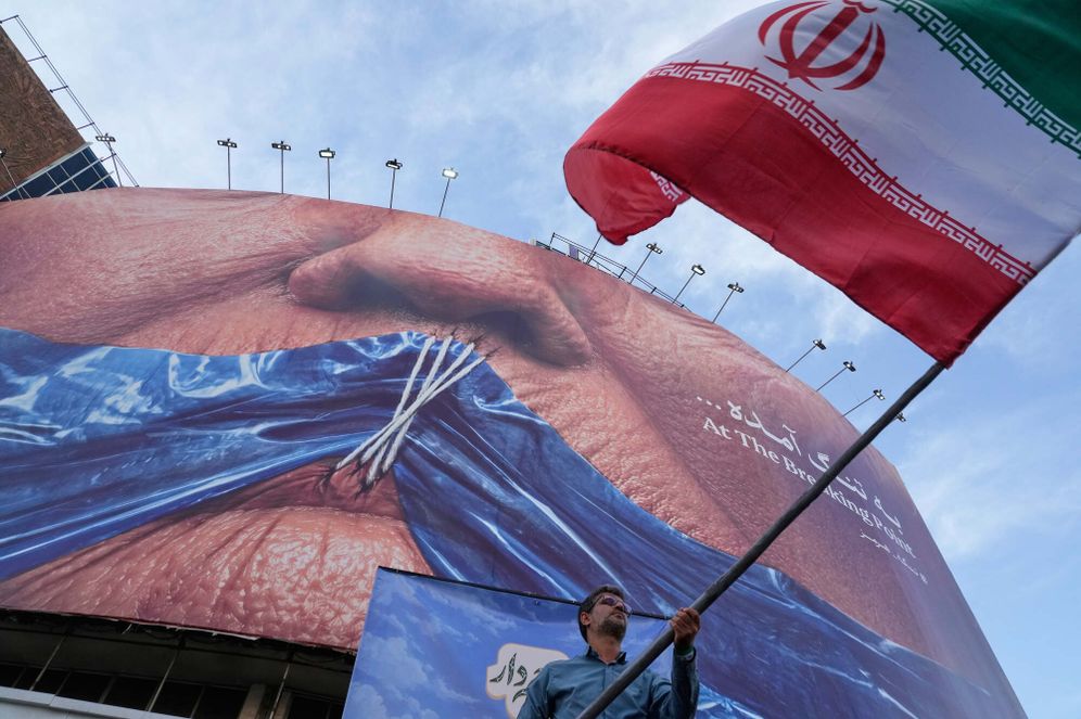 Un homme agite un drapeau iranien pour une campagne progouvernementale sous un panneau d'affichage montrant le détroit d'Ormuz et les lèvres cousues du président américain Donald Trump, sur une place du centre de Téhéran, en Iran, le mercredi 6 mai 2026. 