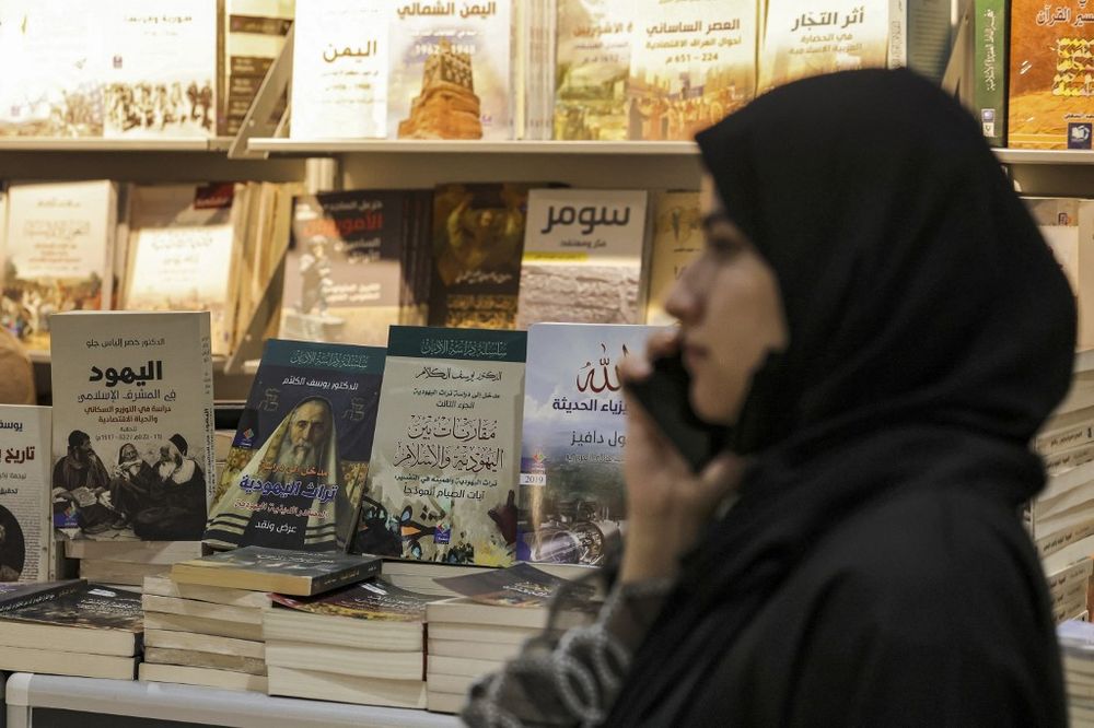 A woman uses her phone as she stands next to a stall displaying Arabic books on Judaism, Islam, Christianity, and comparative religion at the Abu Dhabi International Book Fair in the Emirati capital of the UAE.
