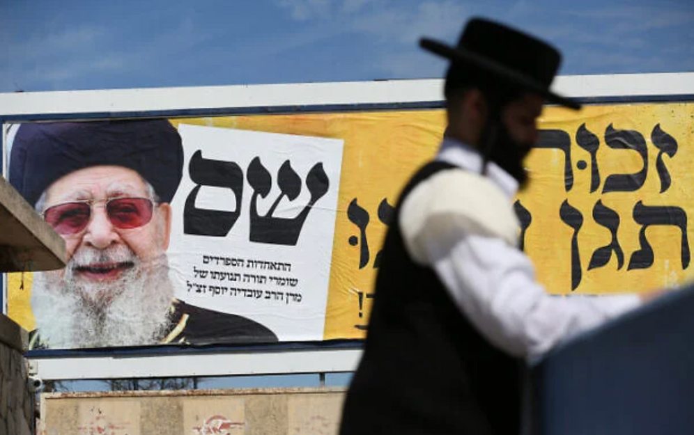 David Cohen/Flash90 Archives : un homme juif orthodoxe passe devant une affiche représentant le défunt rabbin Ovadia Yosef, ancien chef spirituel du Shas, dans le cadre de la campagne électorale du parti, à Safed, dans le nord d’Israël, le 28 février 2020.